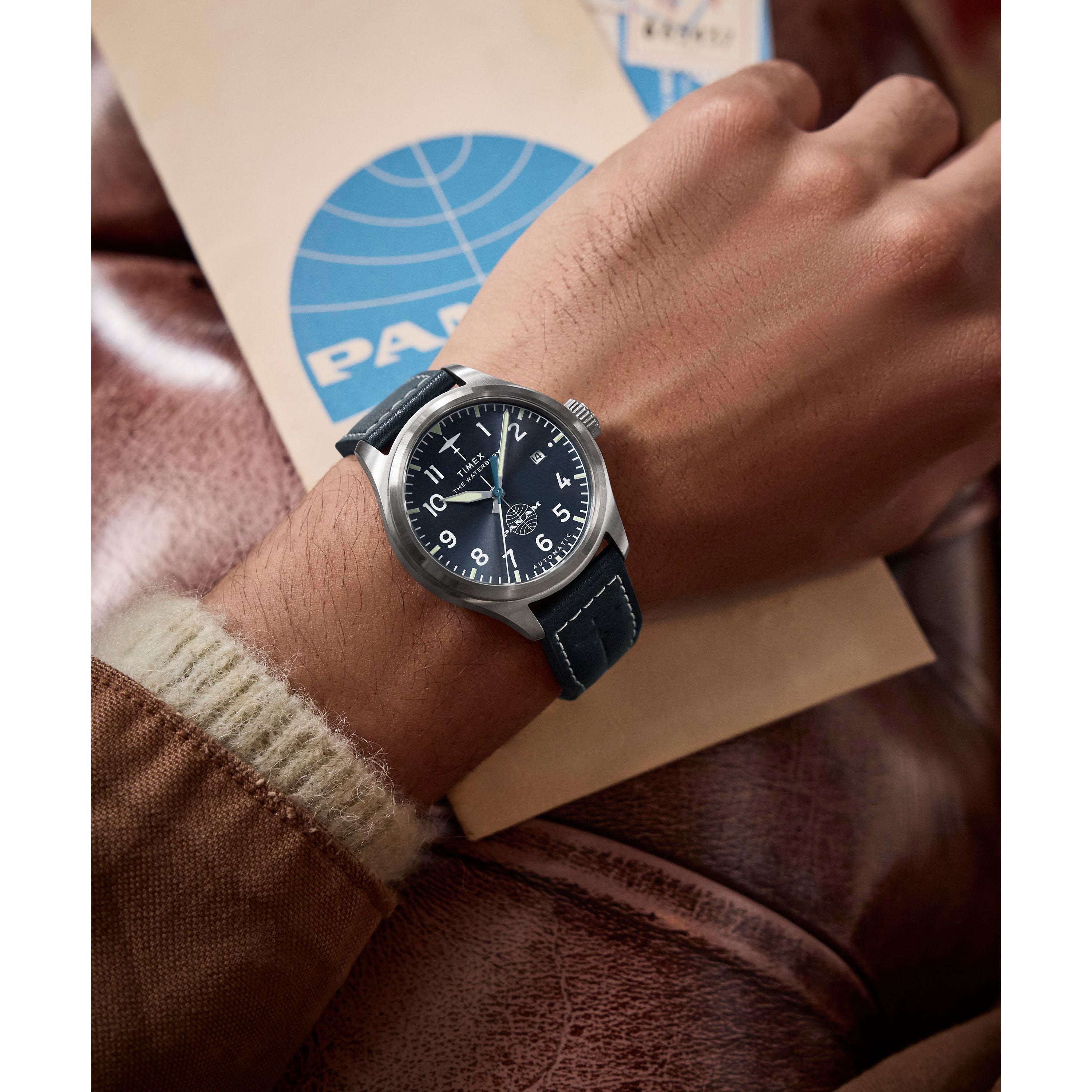 A person wearing a Timex x Pan Am® Waterbury Automatic Ace watch with a black leather strap and stainless steel case rests their hand on a brown leather surface, holding a beige PAN AM envelope with a blue logo.