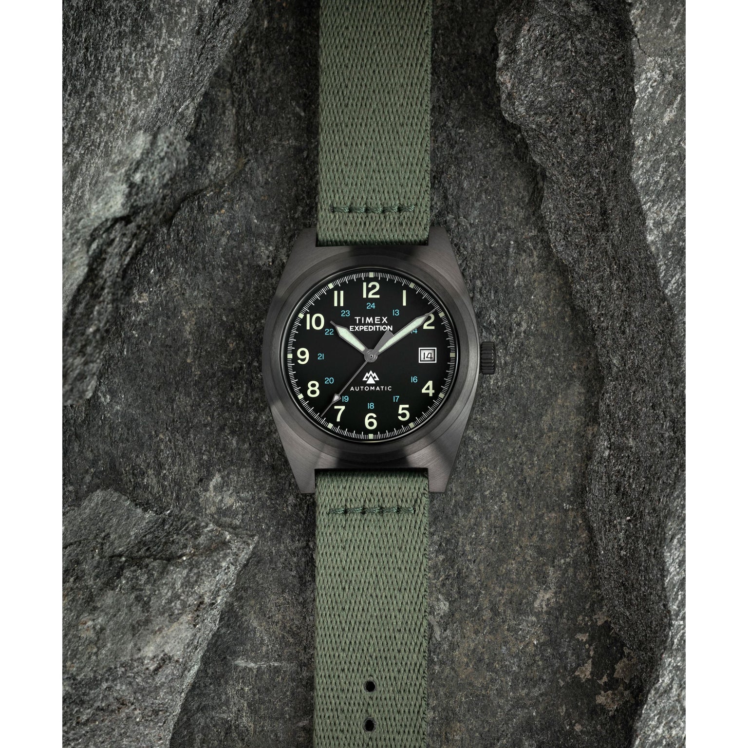 The Timex Expedition® Capstone Automatic wristwatch, with a green fabric strap and black dial with luminous numbers, powered by Japanese automatic movement, is displayed on textured gray rocks.