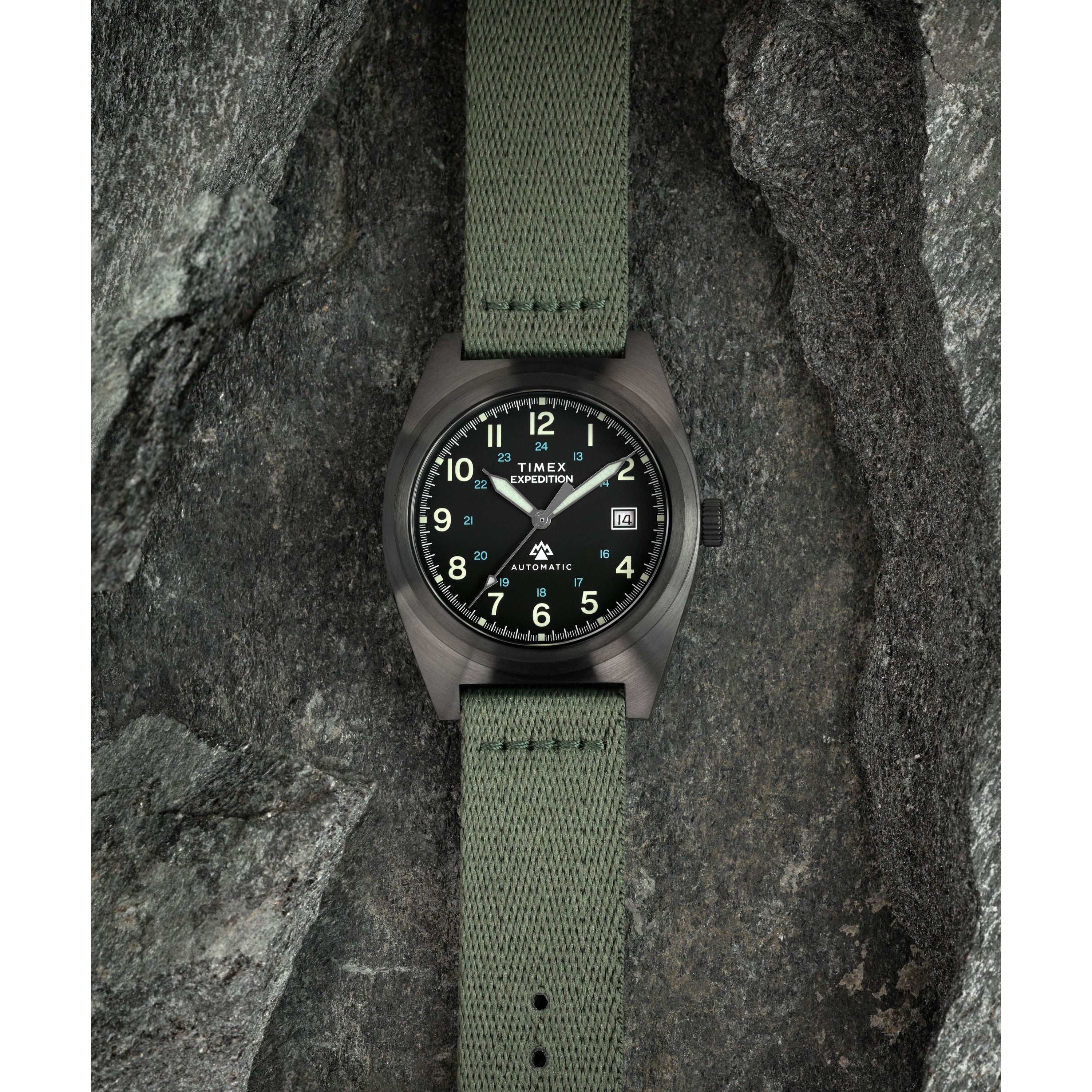 The Timex Expedition® Capstone Automatic wristwatch, with a green fabric strap and black dial with luminous numbers, powered by Japanese automatic movement, is displayed on textured gray rocks.
