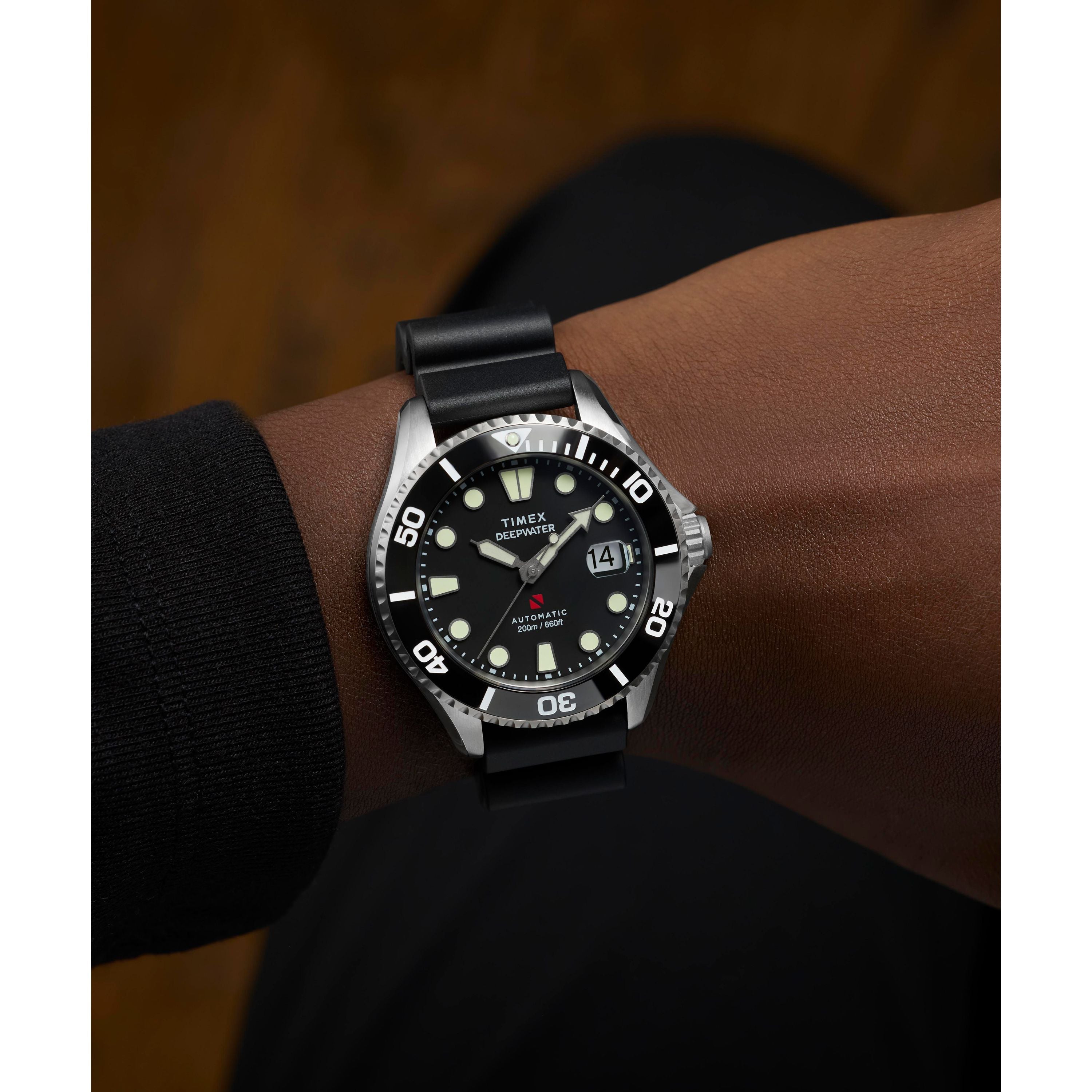A person wears a Timex Deepwater Meridian 200 Automatic dive watch with a black strap, luminous indices, and 200m water resistance. The watch shows 10:10 and the date as the 24th against a brownish blurred background.