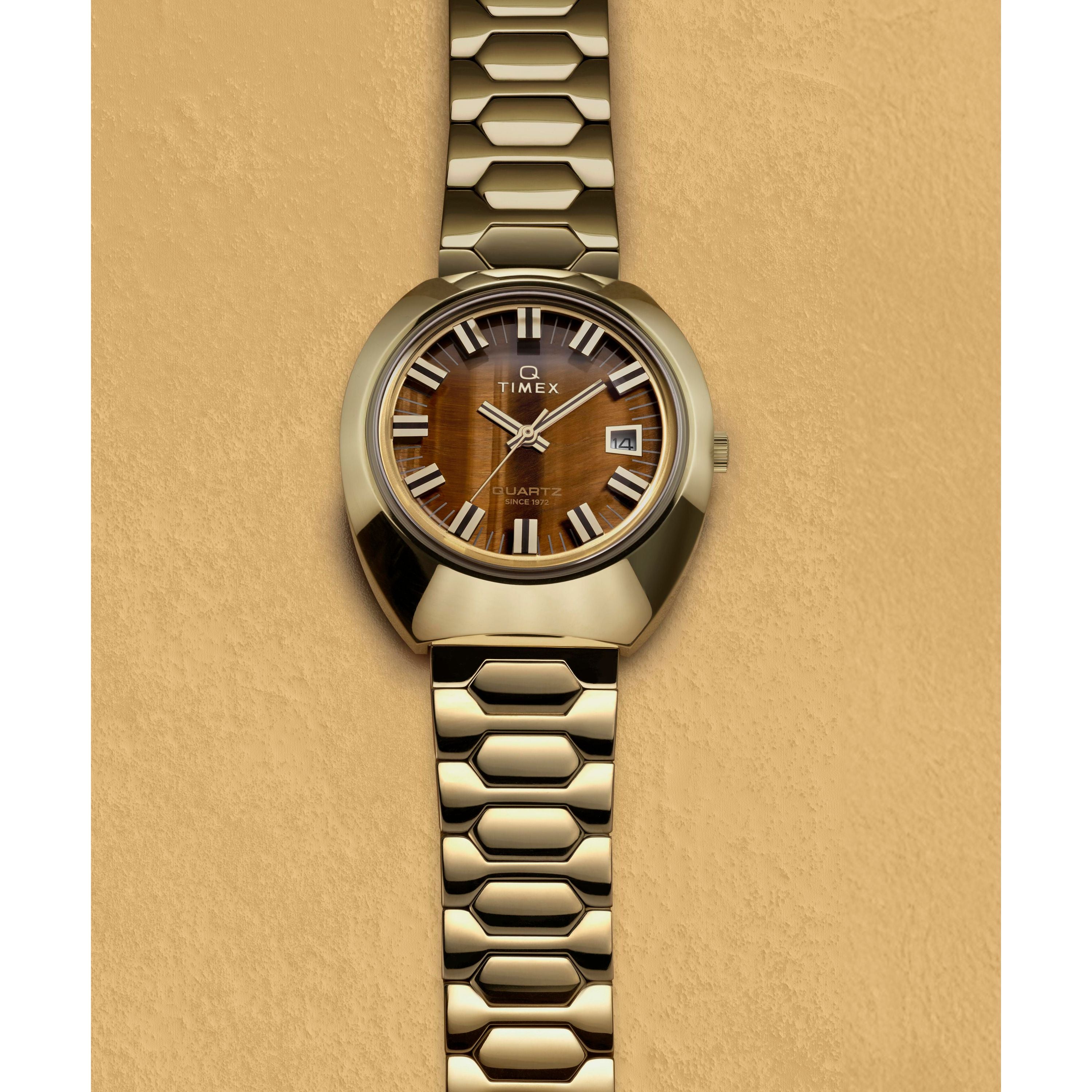 The Timex Q 1972 Time Machine Reissue features a gold-tone case, brown wood-textured dial with bold white markers, and a geometric gold metal bracelet, shown on a tan textured background.
