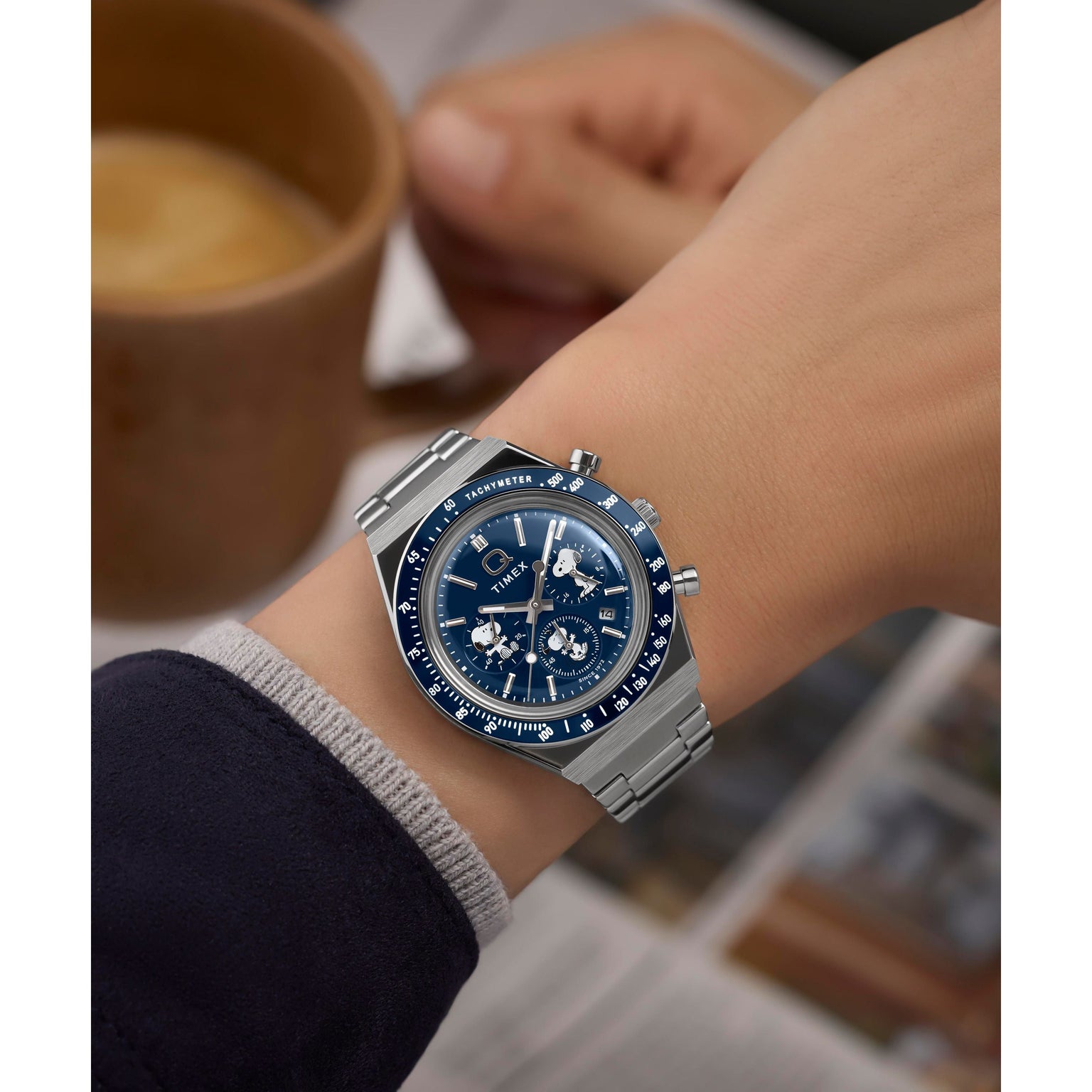 Wearing a Timex x Peanuts® Q Chronograph with a stainless-steel bracelet and blue face, someone holds a cup of coffee as a newspaper sits blurred in the background.