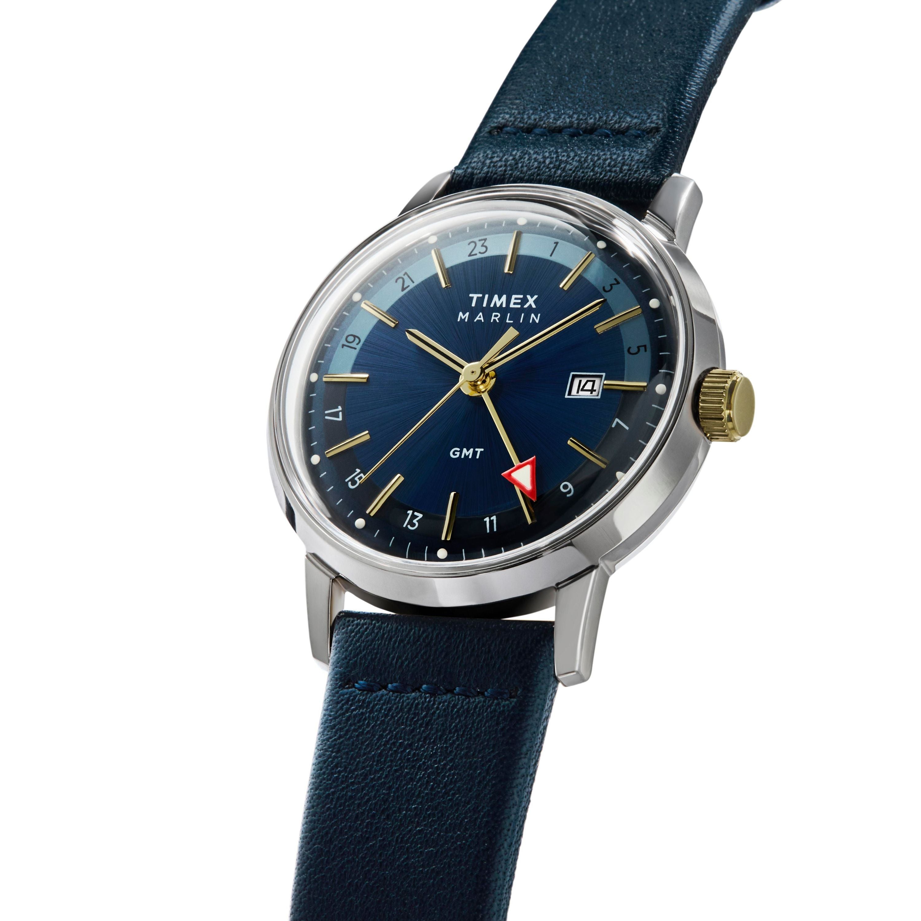The Timex Marlin® Quartz GMT wristwatch features a blue dial with gold accents, a red GMT hand, date window, and blue leather strap with a vintage-style domed acrylic crystal, displayed at an angle on a white background.