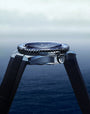A close-up side view of the OMEGA Seamaster Planet Ocean 600M dive watch with a black band, silver bezel, and round face, set against a blurred blue ocean and cloudy sky backdrop.
