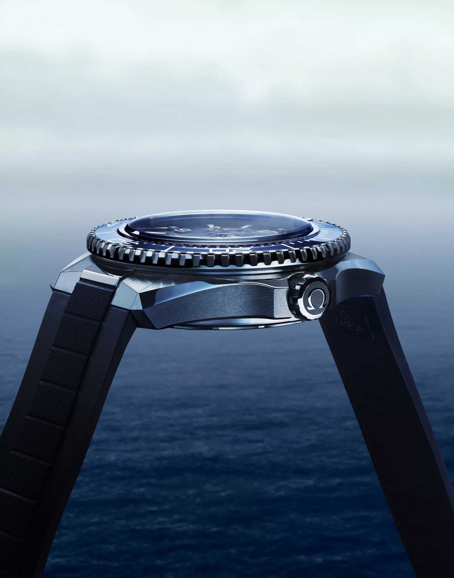 A close-up side view of the OMEGA Seamaster Planet Ocean 600M dive watch with a black band, silver bezel, and round face, set against a blurred blue ocean and cloudy sky backdrop.