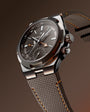 Steel watch with dark dial, dual time subdial, orange accents, and gray fabric strap with orange stitching.