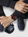 Person wearing a smartwatch with a black case and red display, sitting on a white surface.