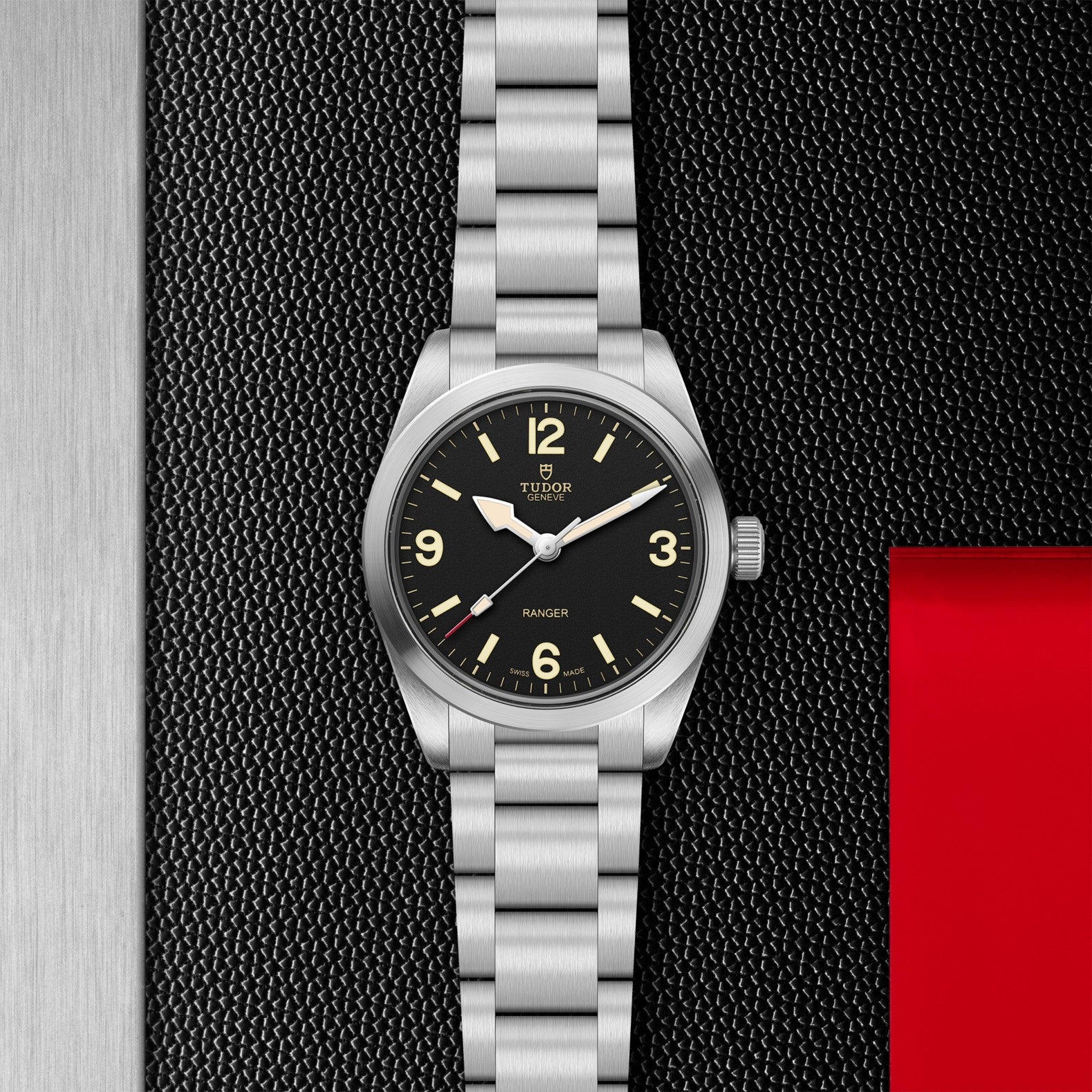 A TUDOR Ranger stainless steel watch with a black dial, luminous hour markers, and brushed metal bracelet is displayed on a textured black surface featuring a red accent on the right.