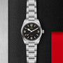 A TUDOR Ranger stainless steel watch with a black dial, luminous hour markers, and brushed metal bracelet is displayed on a textured black surface featuring a red accent on the right.