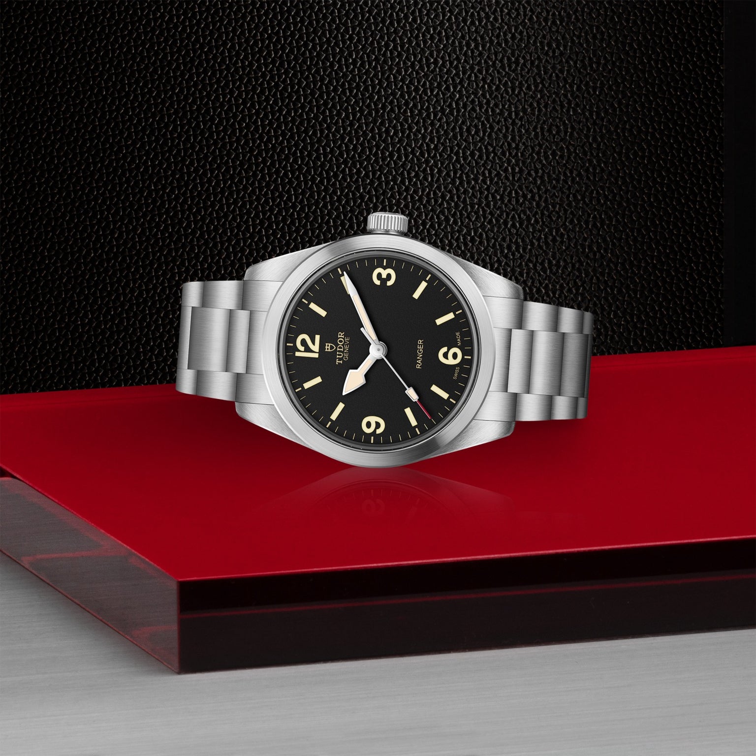 A TUDOR Ranger watch with a stainless steel case, black dial, and luminous hands sits on a red platform against a textured black background, showing 10:10 and featuring an elegantly simple design.
