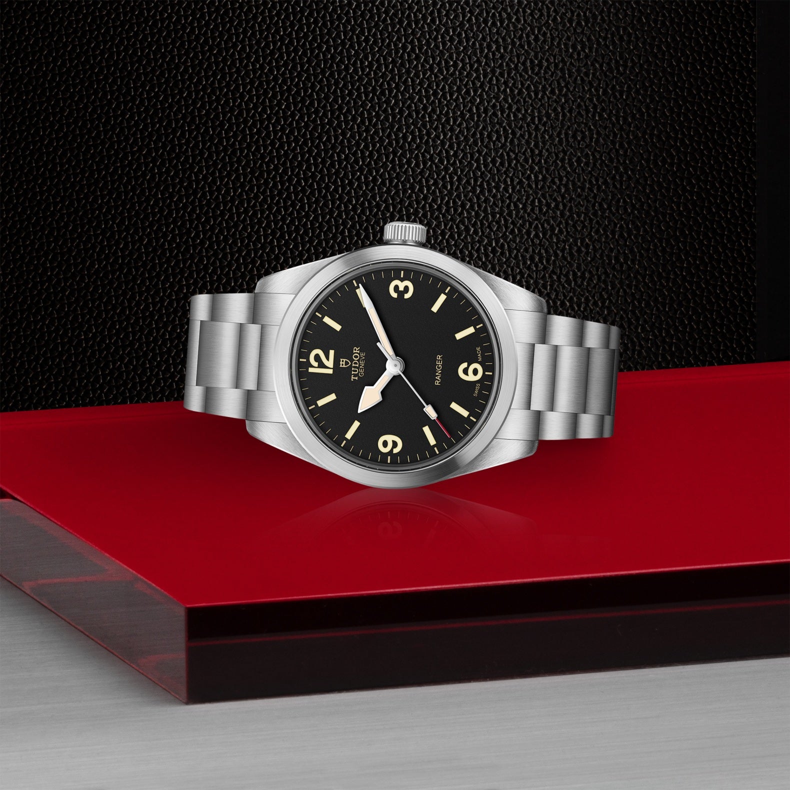 A TUDOR Ranger watch with a stainless steel case, black dial, and luminous hands sits on a red platform against a textured black background, showing 10:10 and featuring an elegantly simple design.