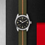 The TUDOR Ranger watch features a stainless steel case, black dial with large cream numerals, and an olive green fabric strap with red and beige stripes, shown on a textured black and silver background with a red accent.