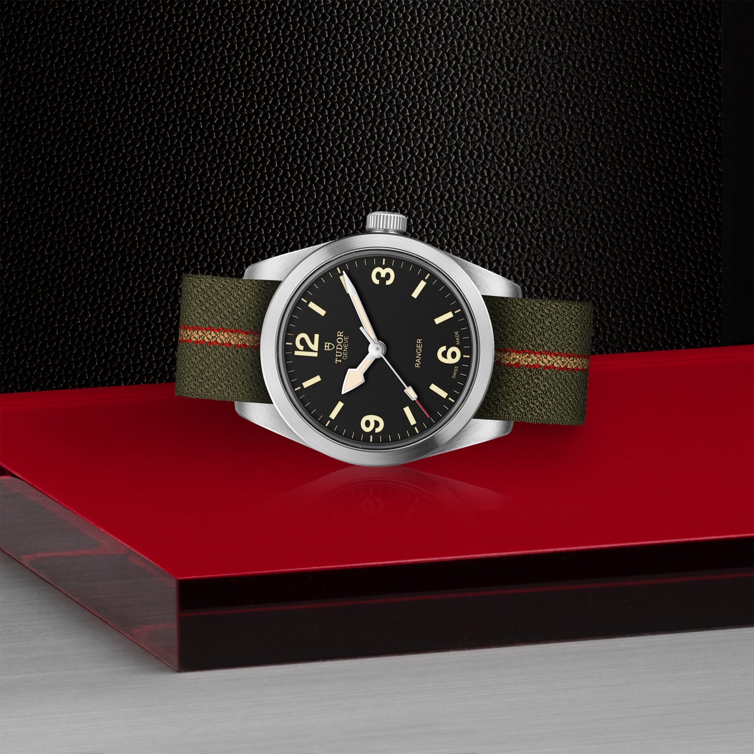 The TUDOR Ranger watch features a black dial, cream markers, and a green fabric strap with an orange stripe, displayed on a glossy red and silver surface against a textured black background.