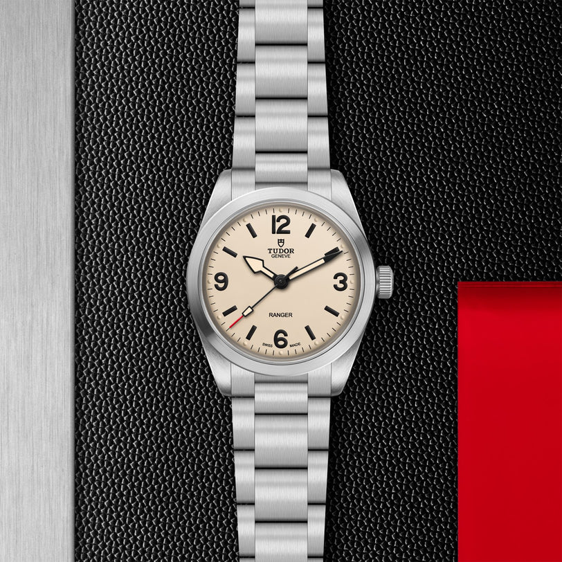 The TUDOR Ranger stainless steel expedition watch features an off-white dial with black numerals and a silver metal bracelet, displayed on a textured black background with metallic and red accents.