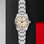 The TUDOR Ranger stainless steel expedition watch features an off-white dial with black numerals and a silver metal bracelet, displayed on a textured black background with metallic and red accents.