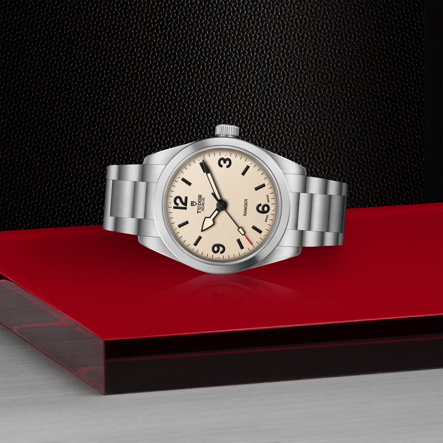 A TUDOR Ranger wristwatch with a cream dial, black numerals, and a stainless steel bracelet rests on a glossy red surface with a textured black background.