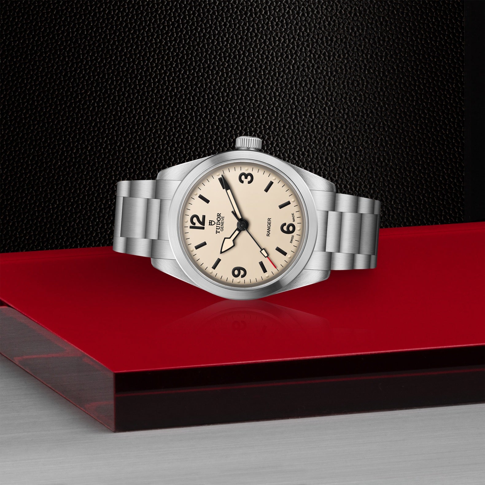 A TUDOR Ranger wristwatch with a cream dial, black numerals, and a stainless steel bracelet rests on a glossy red surface with a textured black background.