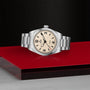 A TUDOR Ranger wristwatch with a cream dial, black numerals, and a stainless steel bracelet rests on a glossy red surface with a textured black background.