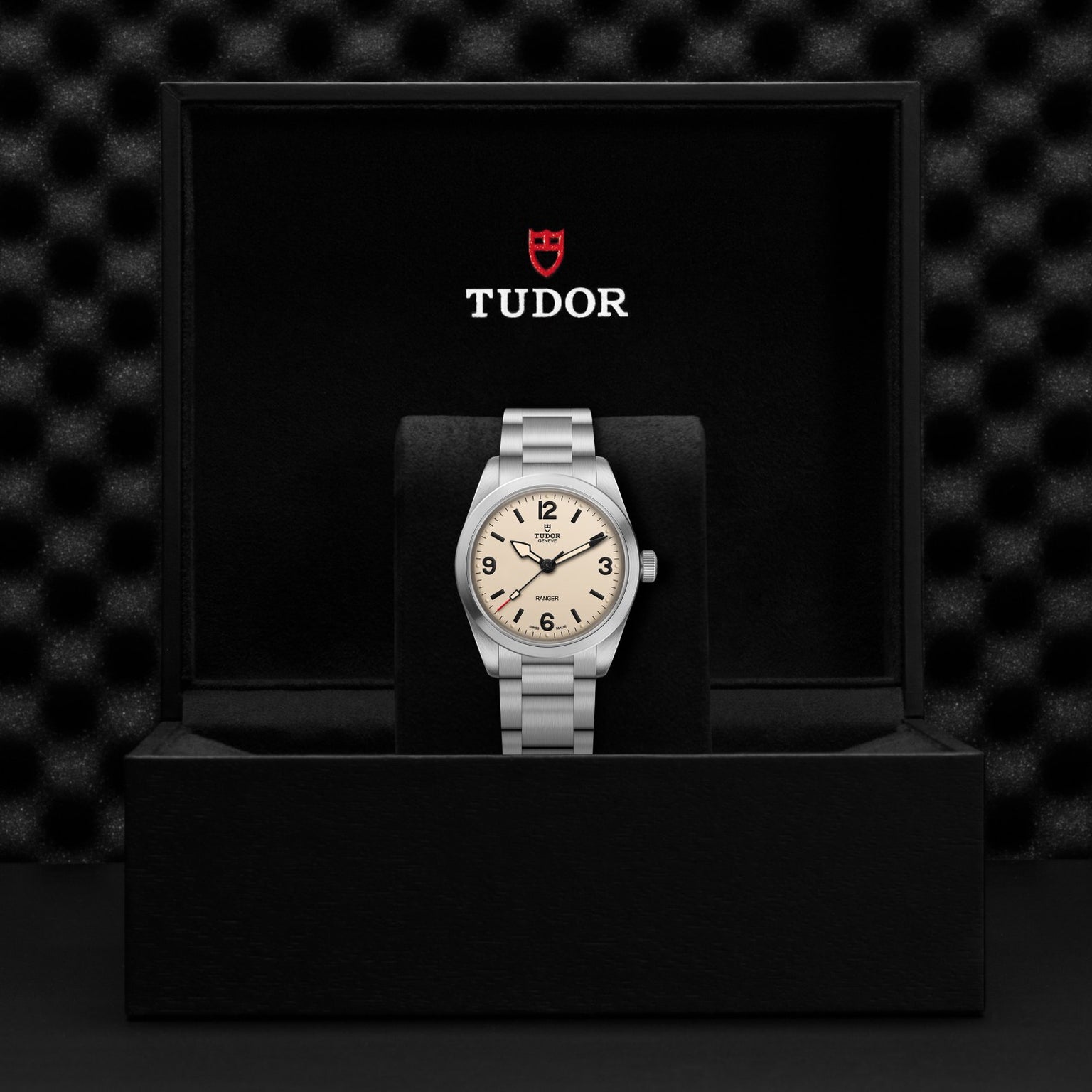 A TUDOR Ranger watch with a light dial is displayed upright in an open black box with the red shield logo above. The stainless steel case contrasts against a textured, dark background.