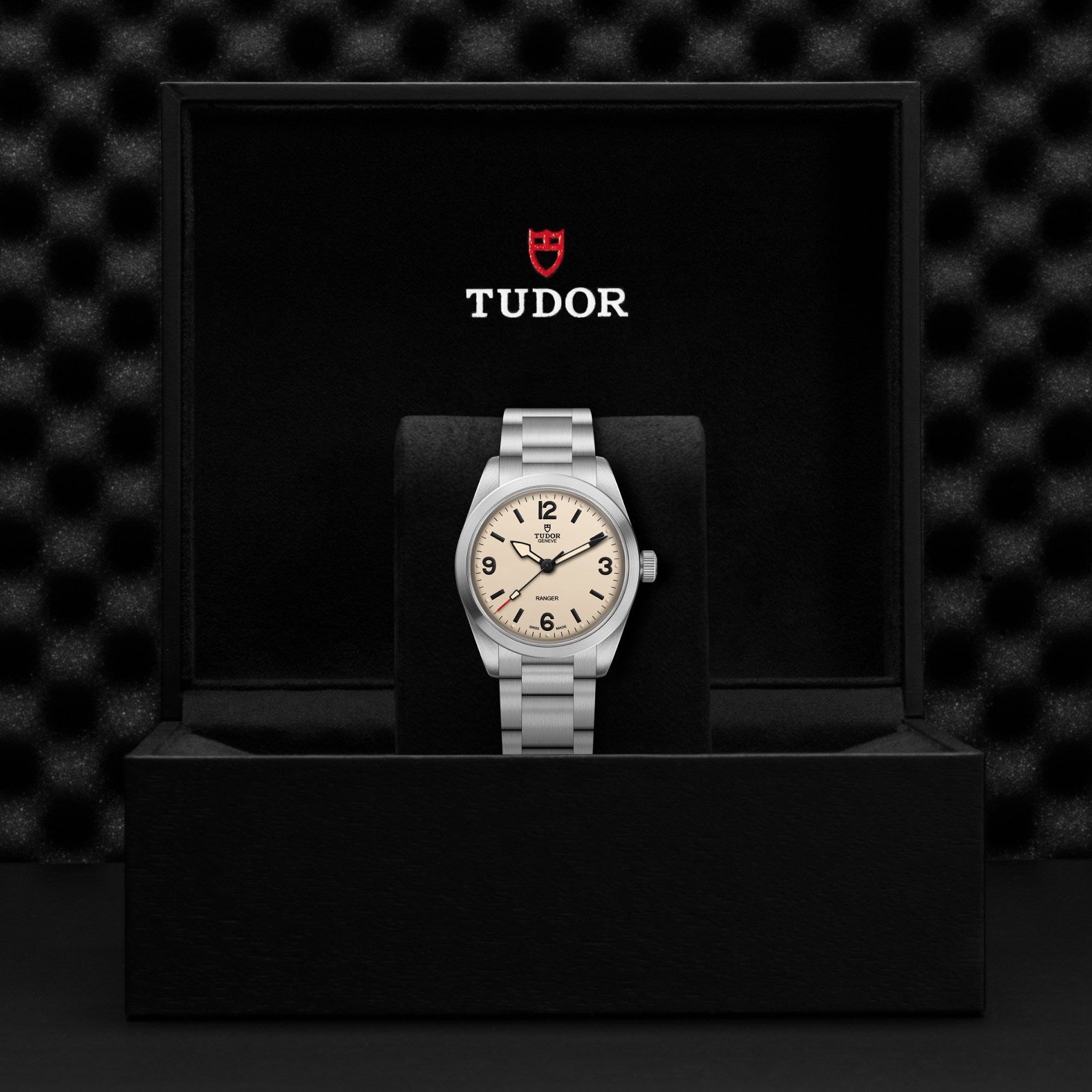 A TUDOR Ranger watch with a light dial is displayed upright in an open black box with the red shield logo above. The stainless steel case contrasts against a textured, dark background.