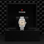 A TUDOR Ranger watch with a light dial is displayed upright in an open black box with the red shield logo above. The stainless steel case contrasts against a textured, dark background.