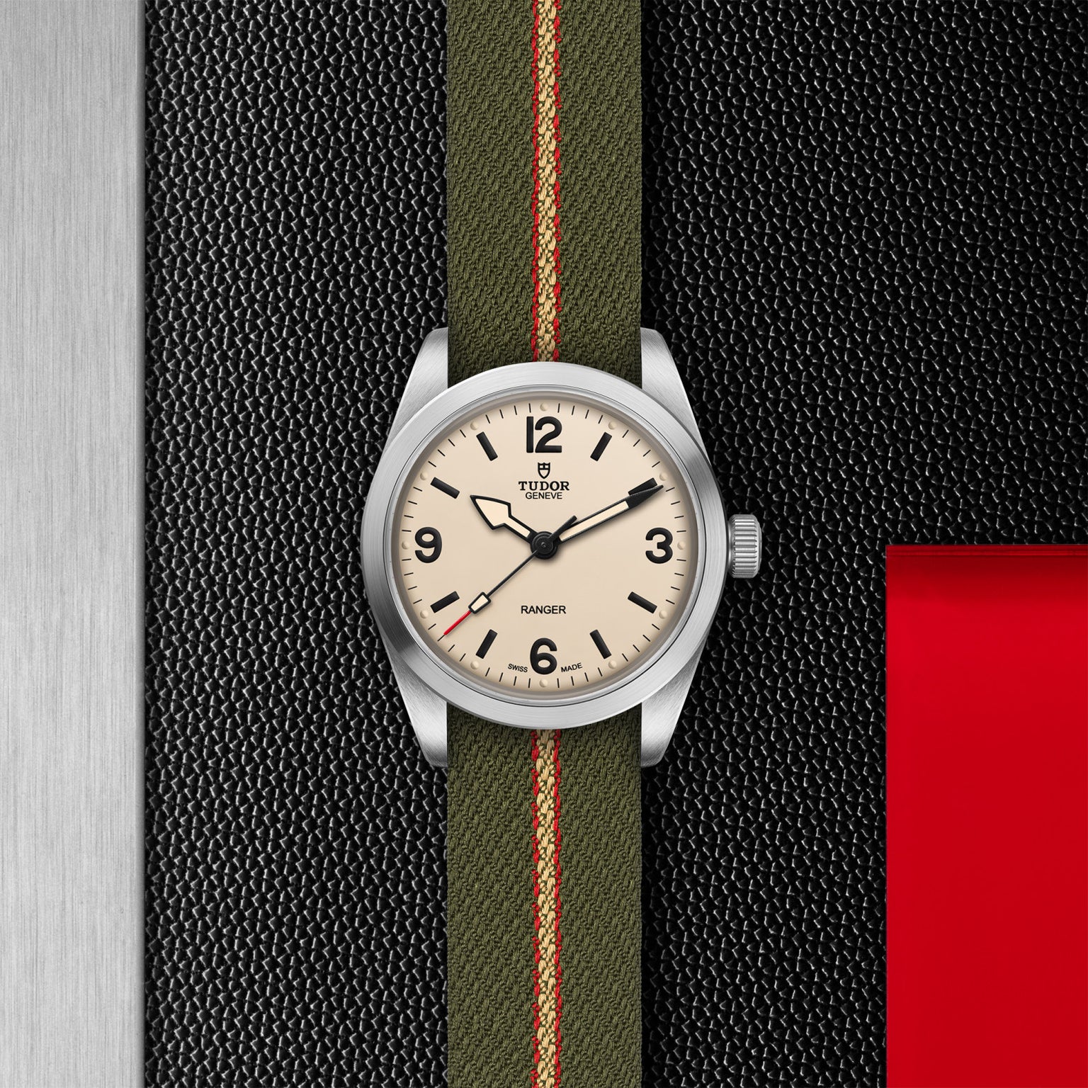 The TUDOR Ranger expedition watch features a cream dial with black numerals and a green fabric strap with red and beige stripes, set against textured black, red, and metallic backgrounds that highlight its robust stainless steel case.