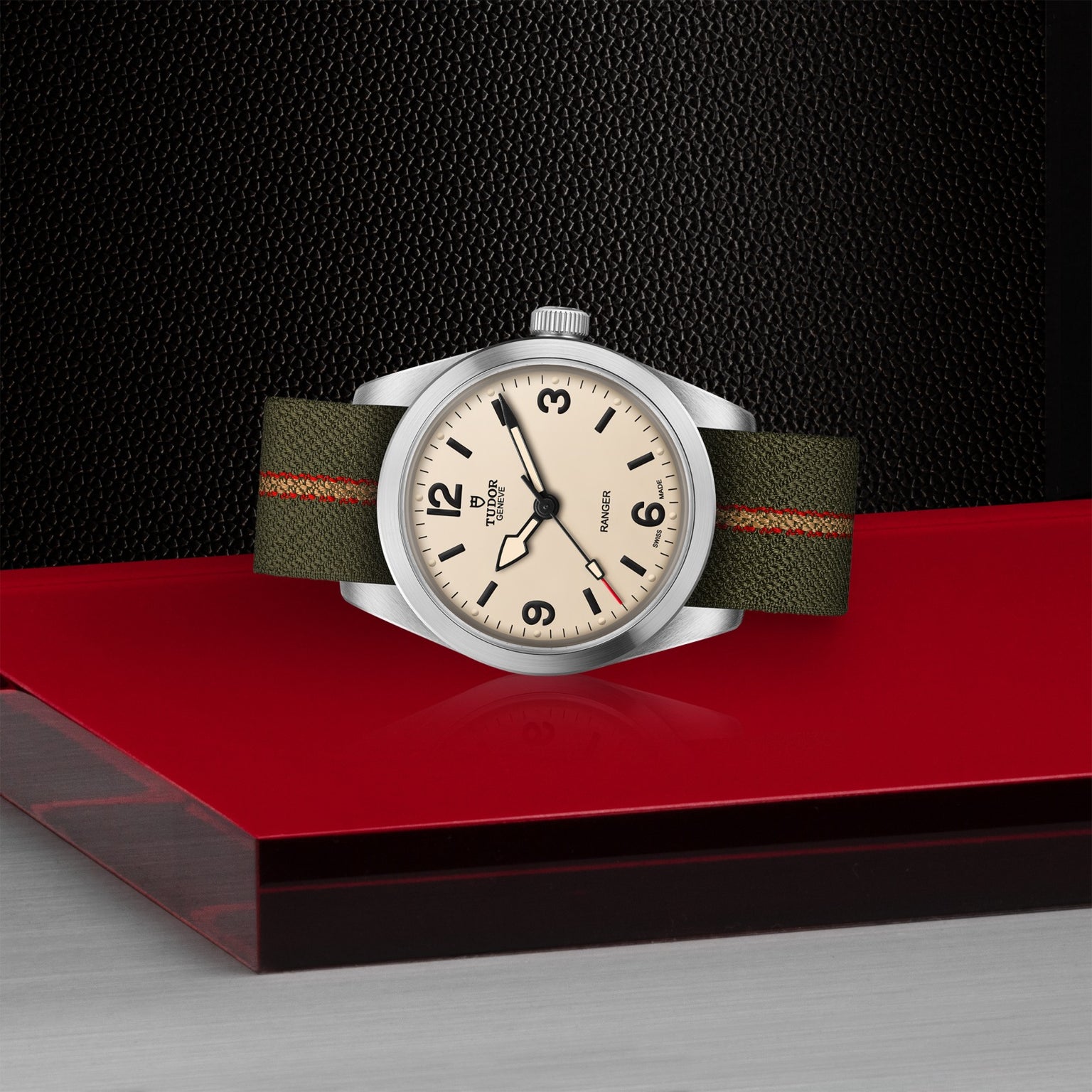 A TUDOR Ranger wristwatch with a beige dial, black numerals, and an olive green fabric strap with an orange stripe is displayed on a glossy red surface against a black textured background.