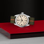 A TUDOR Ranger wristwatch with a beige dial, black numerals, and an olive green fabric strap with an orange stripe is displayed on a glossy red surface against a black textured background.