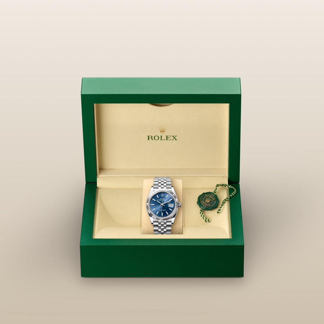 A silver Rolex watch with a blue face in an open green and beige Rolex box with a green tag inside.