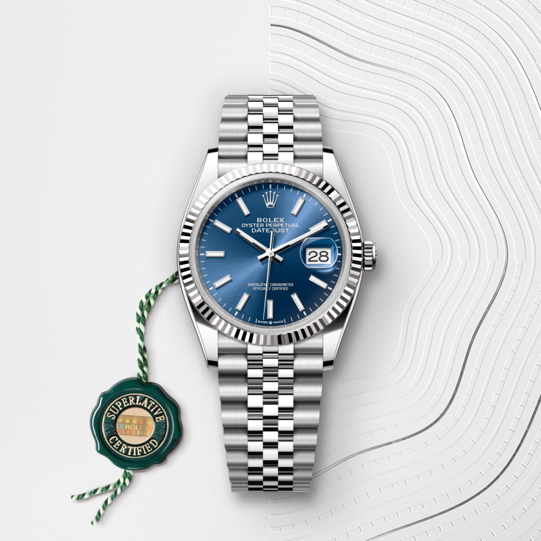 A silver Rolex watch with a blue face and date display, beside a green “Superlative Certified” tag.
