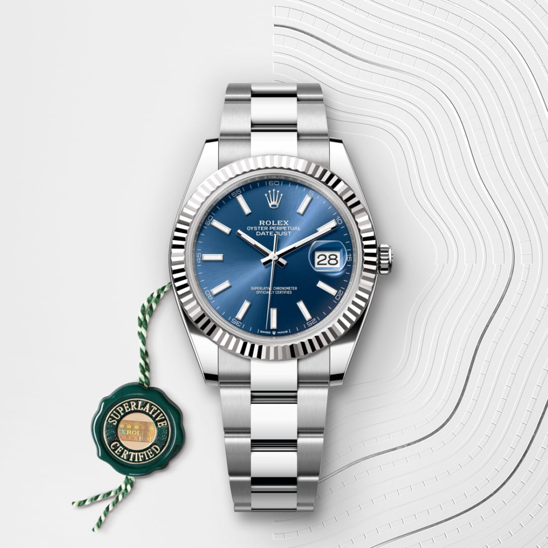 A silver Rolex watch with a blue face and date display, shown on a patterned white background with a green tag.