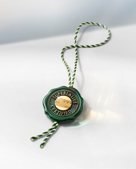 A green and gold Superlative Certified Rolex tag with a twisted green-and-white string on a light surface.