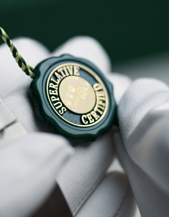 A gloved hand holding a green and gold Rolex Superlative Chronometer certification tag.