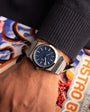 Close-up of a wrist wearing a silver watch with a blue face, against a colorful background.