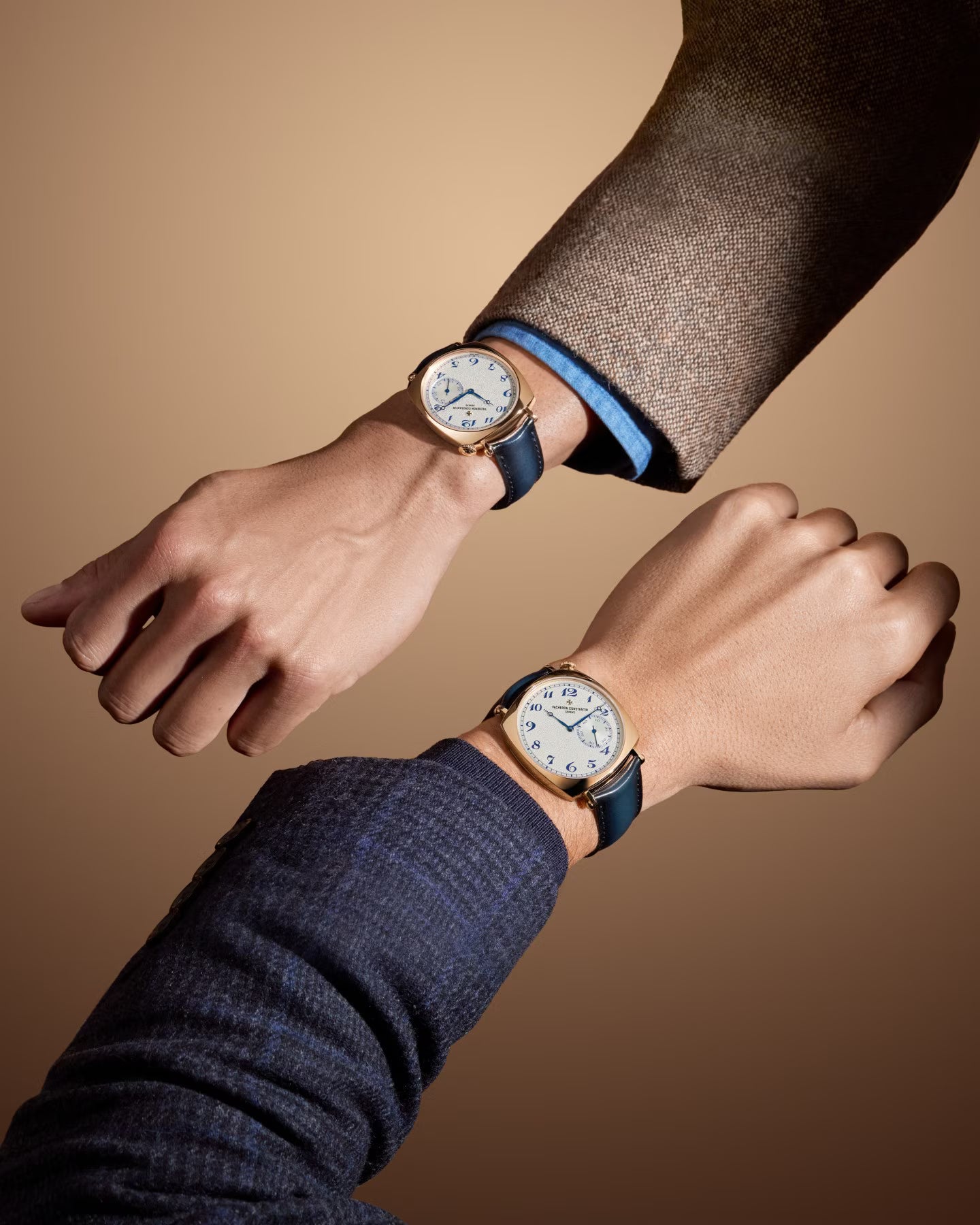 Two wrists wearing round watches with white dials, blue leather straps, and rose gold cases against a neutral tan background.
